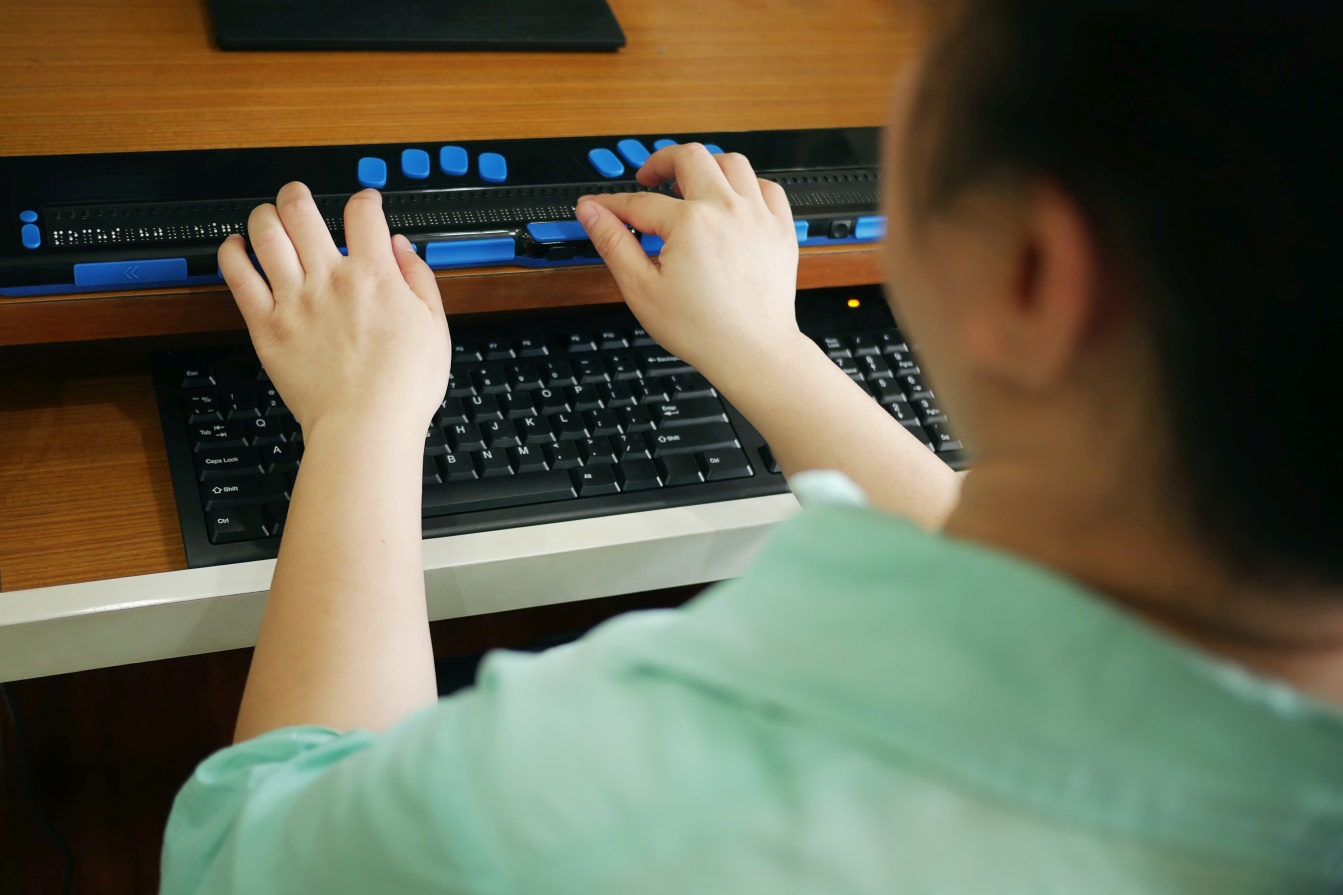 Rückansicht einer Person mit Blindheitsbehinderung, die eine Computertastatur und ein Braille-Display oder ein Braille-Terminal verwendet.